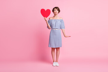 Photo portrait of woman sending air kiss holding big red heart postcard in one hand isolated on pastel pink colored background