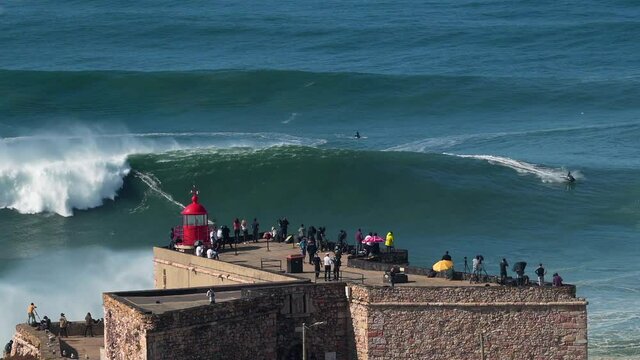 Surfer riding giant wave near historical landmark Fort of Sao Miguel Arcanjo Lighthouse in Nazare, Portugal. Nazare is famously known to surfers for having the largest waves in the world.