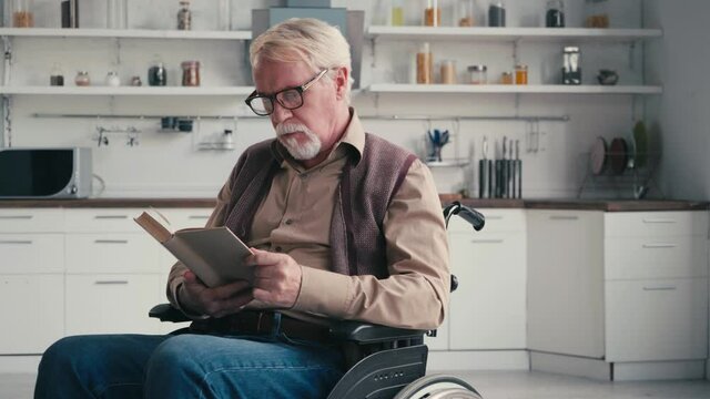 Grey haired man in wheelchair reading book with kitchen on background