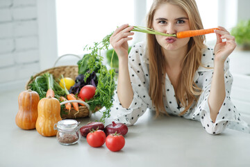 Young and happy woman with carrot at table with green fresh ingredients indoors. High quality photo.
