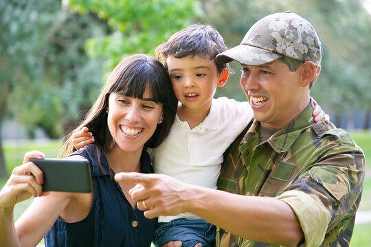 Happy excited military man, his wife and little son taking selfie on cell phone in city park. Front view. Family reunion or returning home concept