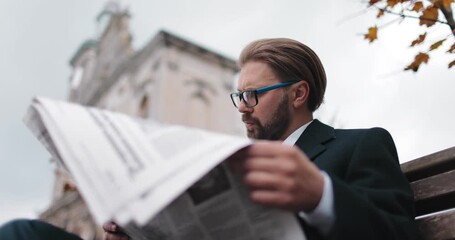 Successful businessman in stylish formal clothing reading daily news while sitting on wooden bench outdoors. Mature man in eyeglasses holding newspaper in hands.
