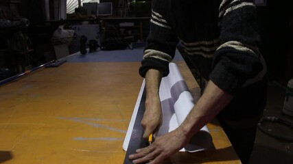 A guy cuts a roll of paper. Preparation of self-adhesive paper for cutting with a conceler knife. The man works in an advertising workshop.
