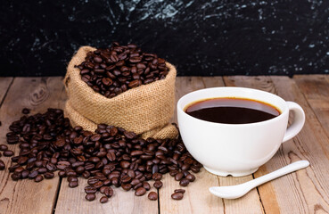 White coffee cup with dark black coffee and a spoon There were fragrant roasted coffee beans in a bag placed on the old wooden floor.