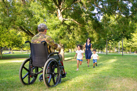 Cheerful Kids And Their Mom Meeting Military Father And Running To Disabled Man In Camouflage. Veteran Of War Or Returning Home Concept