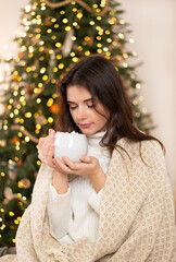 Calm pretty girl sit divan covered checkered blanket have relax in her house with christmas lights illumination hold mug with hot cocoa or coffee. 