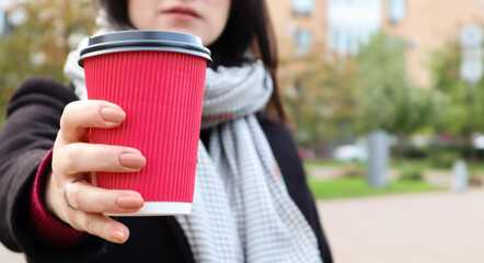 Young stylish woman in a coat and scarf drinks morning hot coffee in a red eco paper glass outdoors...