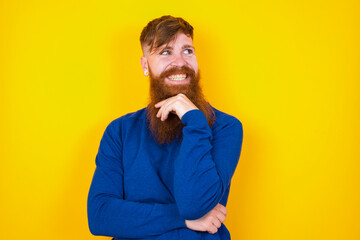 Dreamy Young handsome red haired bearded man standing against yellow wall with pleasant expression, looks sideways, keeps hand under chin, thinks about something pleasant.