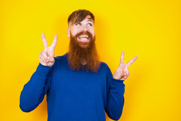 Isolated shot of cheerful Young handsome red haired bearded man standing against yellow wall makes peace or victory sign with both hands, feels cool.