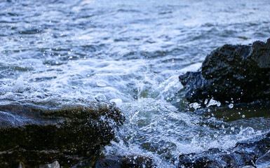 Fototapeta premium waves run onto the shore and crash against the rocks, creating many splashes and splashes near the shore. river surf in stormy weather near a stone pebble coast with foamy splashing waves.