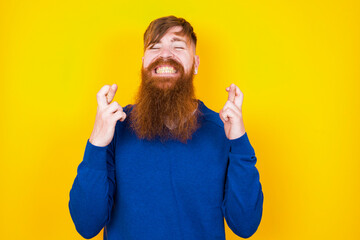 Young handsome red haired bearded man standing against yellow wall holding fingers crossed with...