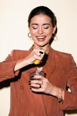 happy woman in brown blazer squeezing lemon in glass with alcohol cocktail on white - obrazy, fototapety, plakaty