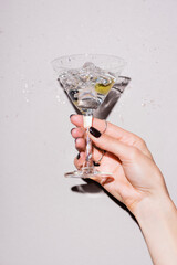 cropped view of woman shaking glass of martini with olive near drops of alcohol on white - obrazy, fototapety, plakaty