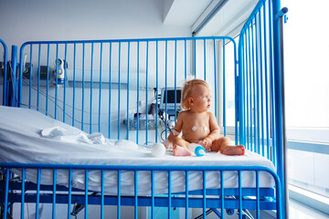 Little toddler sit in hospital bed with catheter in his hand and milk bottle nearby look aside