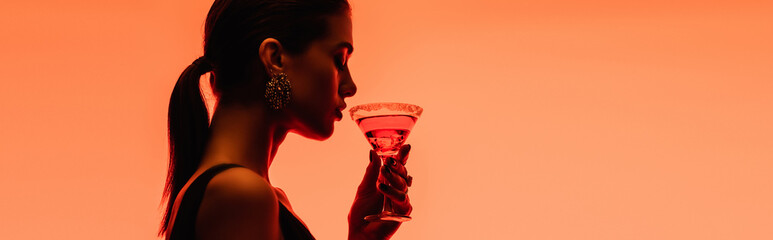 side view of young woman holding margarita cocktail with ice cubes on orange, banner