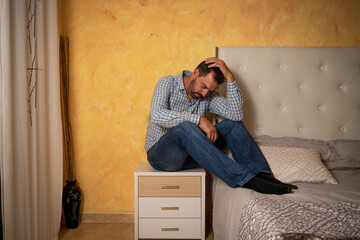 Attractive bearded man depressed and upset in the bedroom. Dramatic 30-40 year old lifestyle portrait, handsome man sitting on bed, worried and desperate
