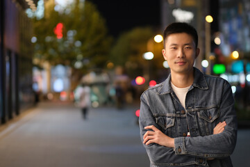 mid shot of one Asian young man at modern city at night, crossed arm and smiling, looking at camera