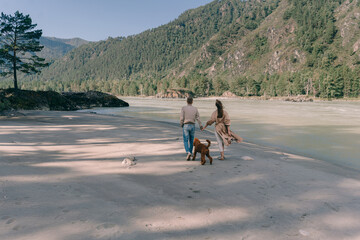 A couple in love with a red poodle walk along the sandy bank of the river against the background of mountains at sunset in Altai