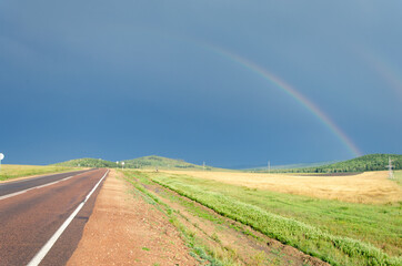 View of the road and rainbow in the sky. On the side of the road fields with green grass and forest.