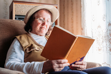 Elderly woman with a hat on, reading a book by the window. Afternoon light