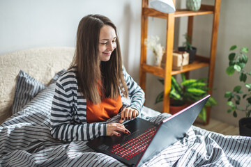 Cute young woman is sitting on a bed at home and using a laptop. Concept of distance learning, remote work, online communication