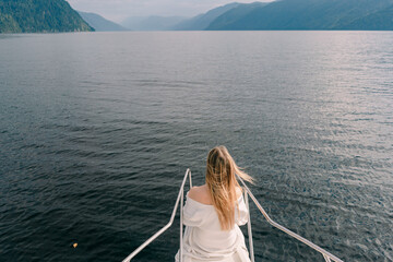 Fototapeta premium A girl with hair fluttering in the wind stands on the bow of a boat floating on Lake Teletskoye against the backdrop of mountains in Altai