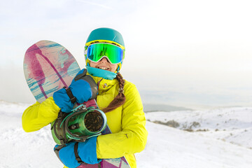 Girl in yellow jacket with a snowboard posing at the panorama of the mountains.