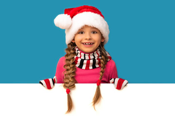 A happy little girl looks out from behind a white background in a Christmas hat.