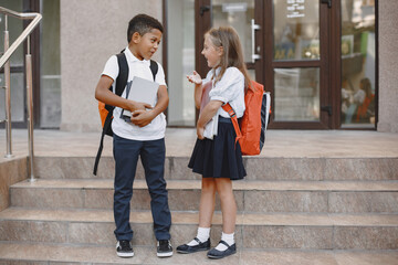 Mixed-races boy and little girl. Junior students in front of school. Pretty Caucasian girl. African...