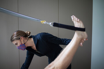 Female pilates instructor wearing mask assisting unrecognizable man on reformer