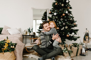 Merry Christmas and Happy Holidays. Cheerful father hugging cute baby daughter girl near Christmas tree. Dad and little child having fun and playing together at home.