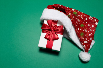 Top view of white gift box with red bow wearing Santa hat on background of green color with copy space.