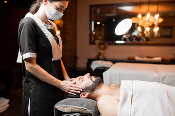 Professional female masseur in medical mask doing facial massage to a male client at Spa salon....