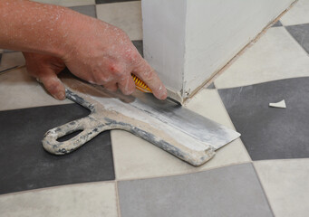 A building contractor installing linoleum flooring near the drywall partition wall is cutting along the crease of black and white linoleum using a sharp utility knife and a putty knife.