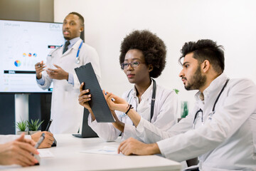 Close up portrait of two young doctors, African woman and Indian man, sitting at the table and discussing medical case. Handsome African male doctor having speech on the background