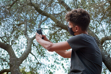 Young man sitting on a tree taking a selfie wearing a face mask-concept of the new normal