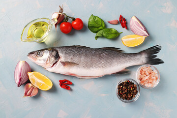 Fresh ready to cook raw seabass with ingredients and seasonings like basil, lemon, salt, pepper, cherry tomatoes and garlic on light blue background. View from above