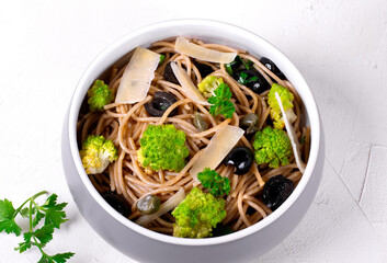 Pasta salad with whole grain spaghetti, romanesco cauliflower, olives, capers and parmigiano cheese in the grey ceramic bowl on the white wooden table