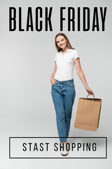 joyful young woman holding shopping bag while standing with hand in pocket near black friday...