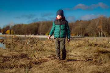 Naklejka na ściany i meble 6 year old caucasian boy wearing warm jacket and knit beanie waling along overgrown shore of finland gulf in Russia holding a stick in hand squinting into the sun, on sunny autumn day 