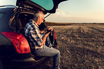 Senior farmer siting in car boot in the field, looking at phone after wheat harvest at sunset.