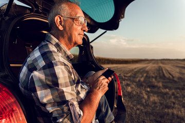 Senior farmer siting in car boot, watching wheat field after harvest at sunset.