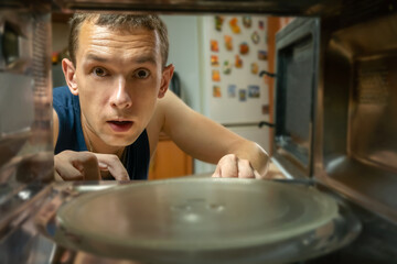 Dark-haired man has opened the microwave oven and looks at it with a surprised look, a photo from inside.
