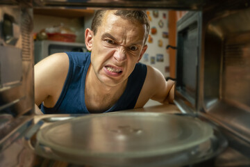 Dark-haired man has opened an empty microwave oven and is looking at it with an angry look, a photo from inside.