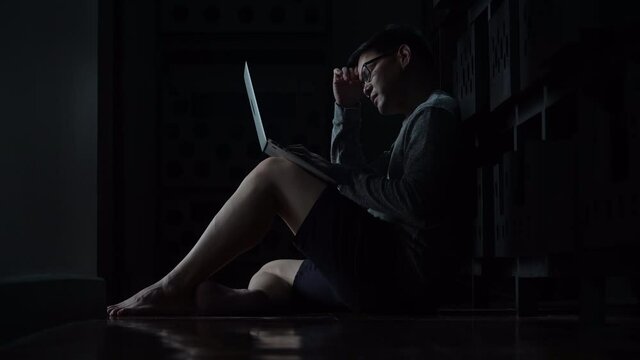 Worried young asian man sitting on the floor with laptop computerin  the dark at home