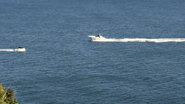 A motorboat in slow-mo sails the waves of a calm sea, encounters a smaller boat travelling in the other direction, then almost disappears behind the cliff. Shot at high frame rate to slow speed down.