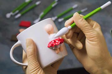 A master makes a figurine of a person on a ceramic mug made of polymer clay, close-up