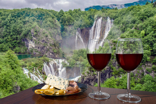 Two glasses of wine with meat and cheese snacks against beautiful big waterfalls in forest in Croatia. Glass of red wine with different snacks - plate with ham, sliced, blue cheese.