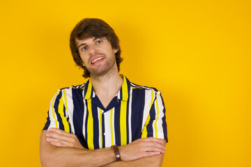 Young handsome Caucasian man, wearing stripped shirt standing against yellow wall being happy smiling and crossed arms looking confident at the camera. Positive and confident person.