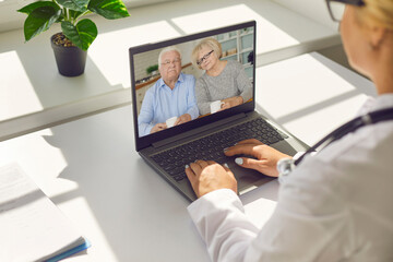 Faceless doctor sitting in her office and talking to a senior couple via video call using a laptop.